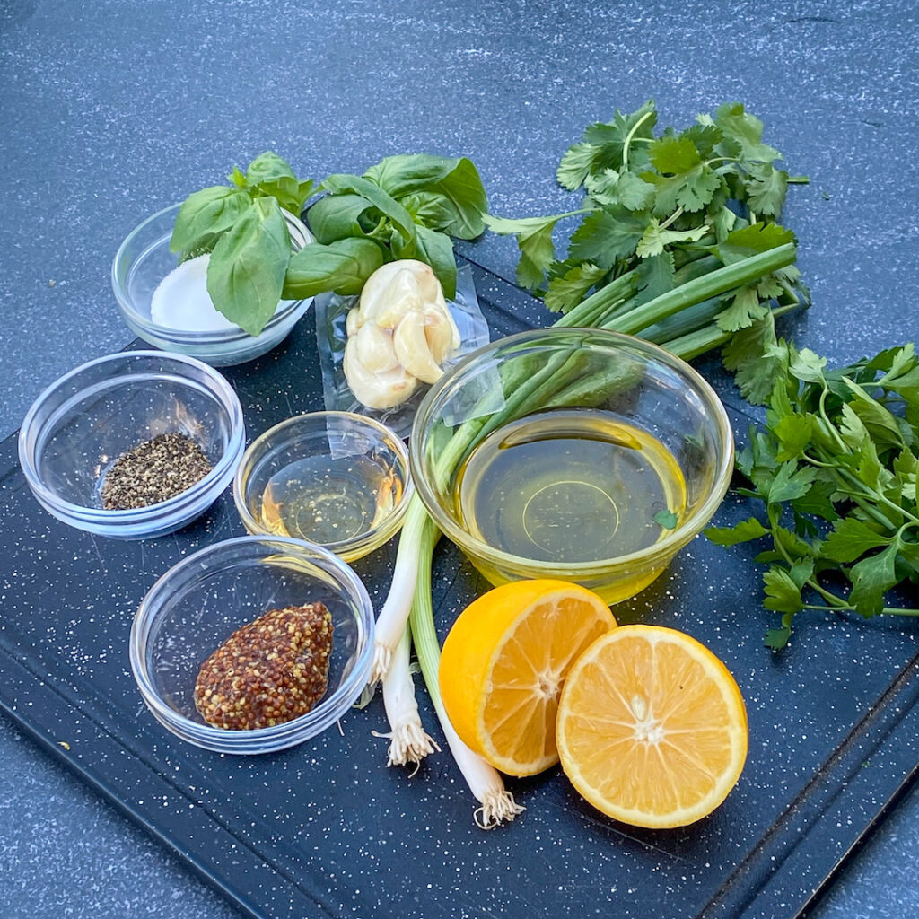 On a chopping block the ingredients for the herb marinade include, Meyer Lemon, whole grain mustard, oil, agave, pepper, green onions, basil, parsley, cilantro, and garlic.  
