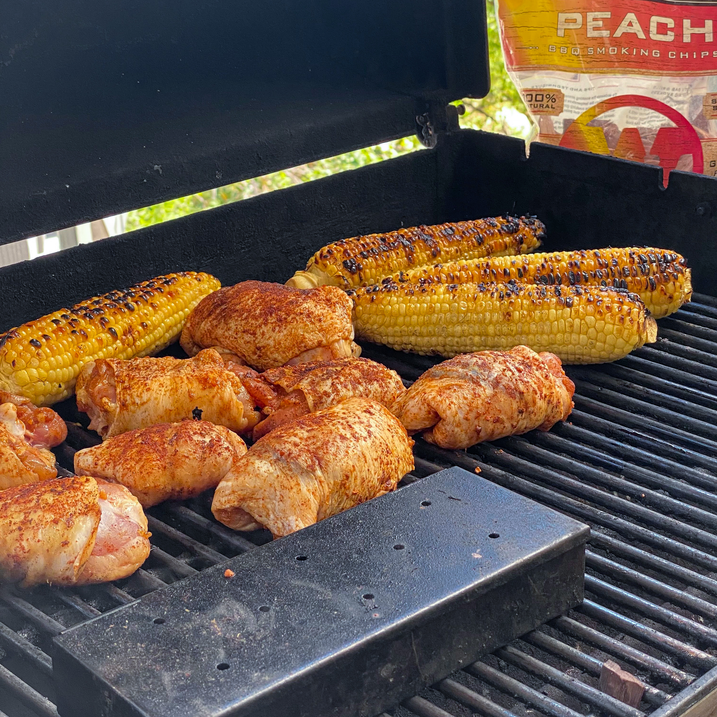 On the grill, a smoker box that is filled with peach smoking chips with prepared chicken thighs ready to cook. 