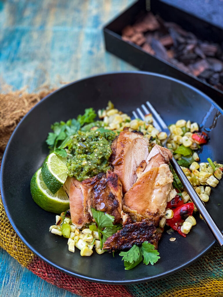 An eye-appealing plate of sliced smoked chicken, smoked salsa verde, and charred corn salad. Garnishes include lime slices and cilantro. 