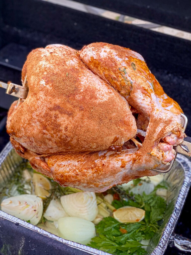 Seasoned turkey has just been put on a rotiserrie grill. Aeromatics are in a pan below.