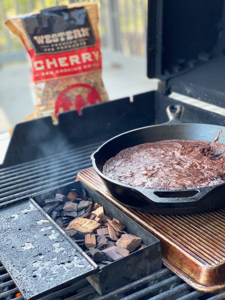 An open grill with smoking chips, a brownie in a skillet, and a bag of Western Cherry BBQ Smoking Chips. 