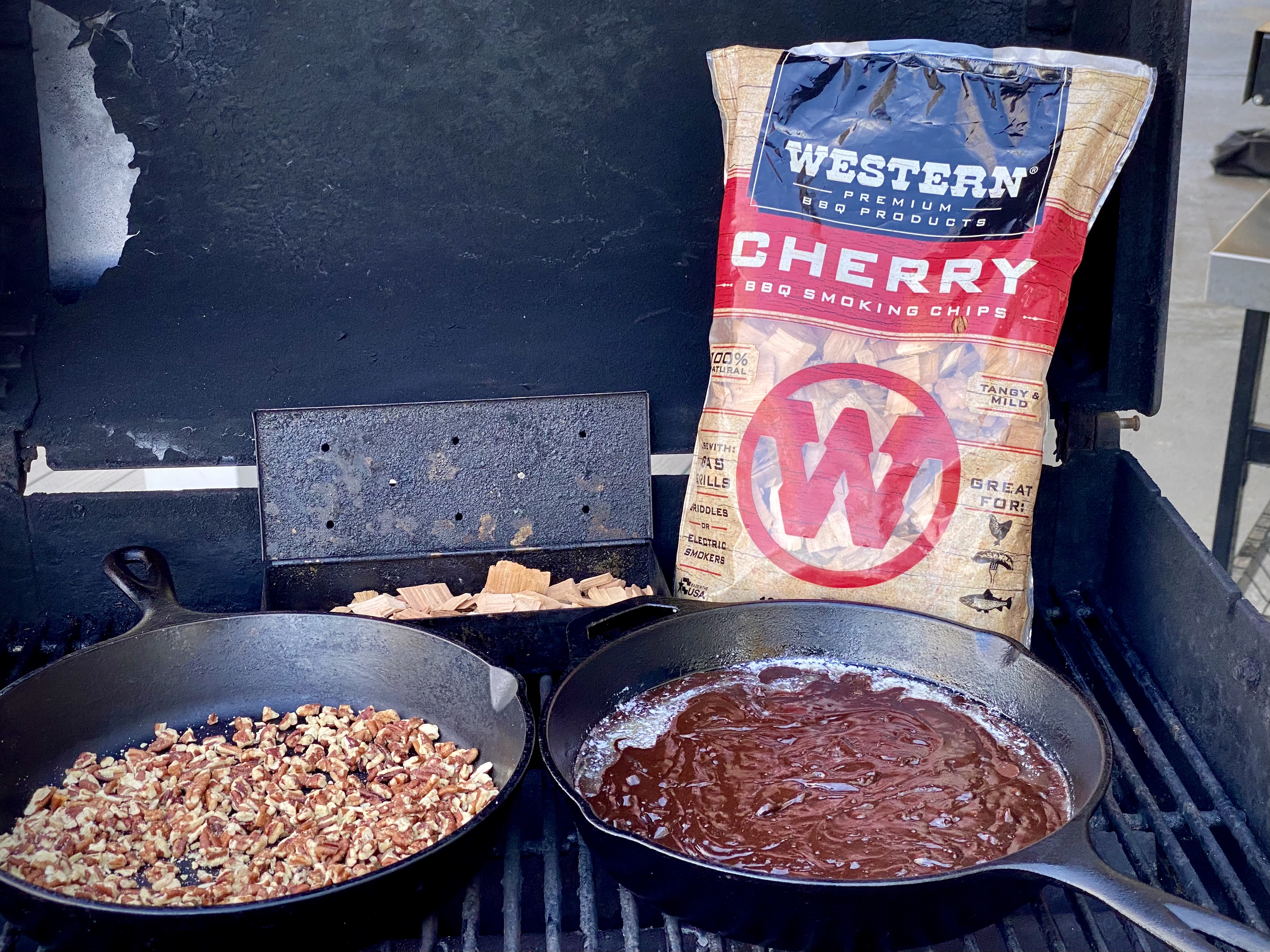 A bag of Western Cherry BBQ Smoking Chips on a grill with a skillet of pecans and a skillet of brownie. 