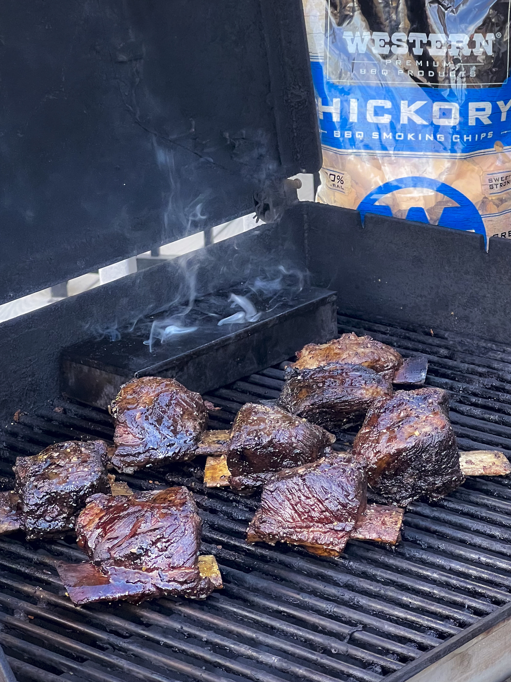 Short Ribs on a grill with a smoke drifting from a smoker box. A bag of Western Hickory Chips is in backdrop.