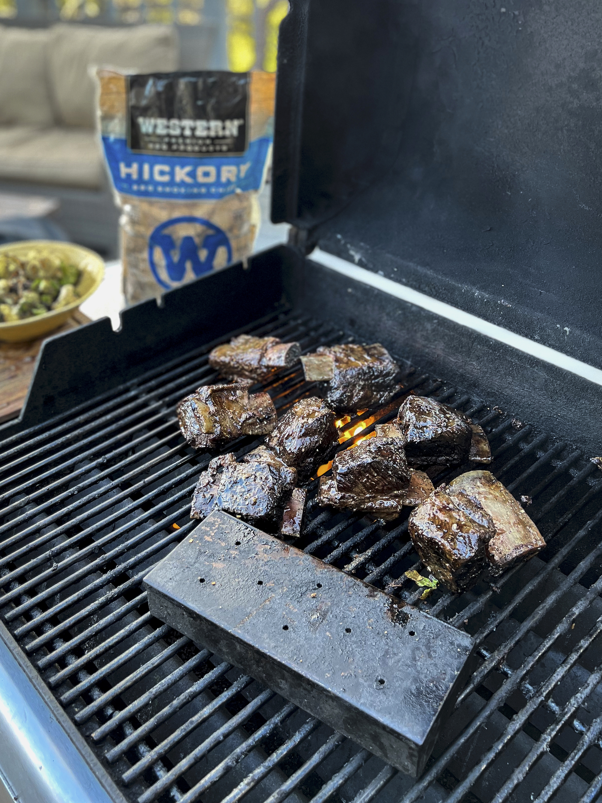 A smoker box on the grill with partially cooked meat on bones. A bag of hickory smoking chips is in the background. 