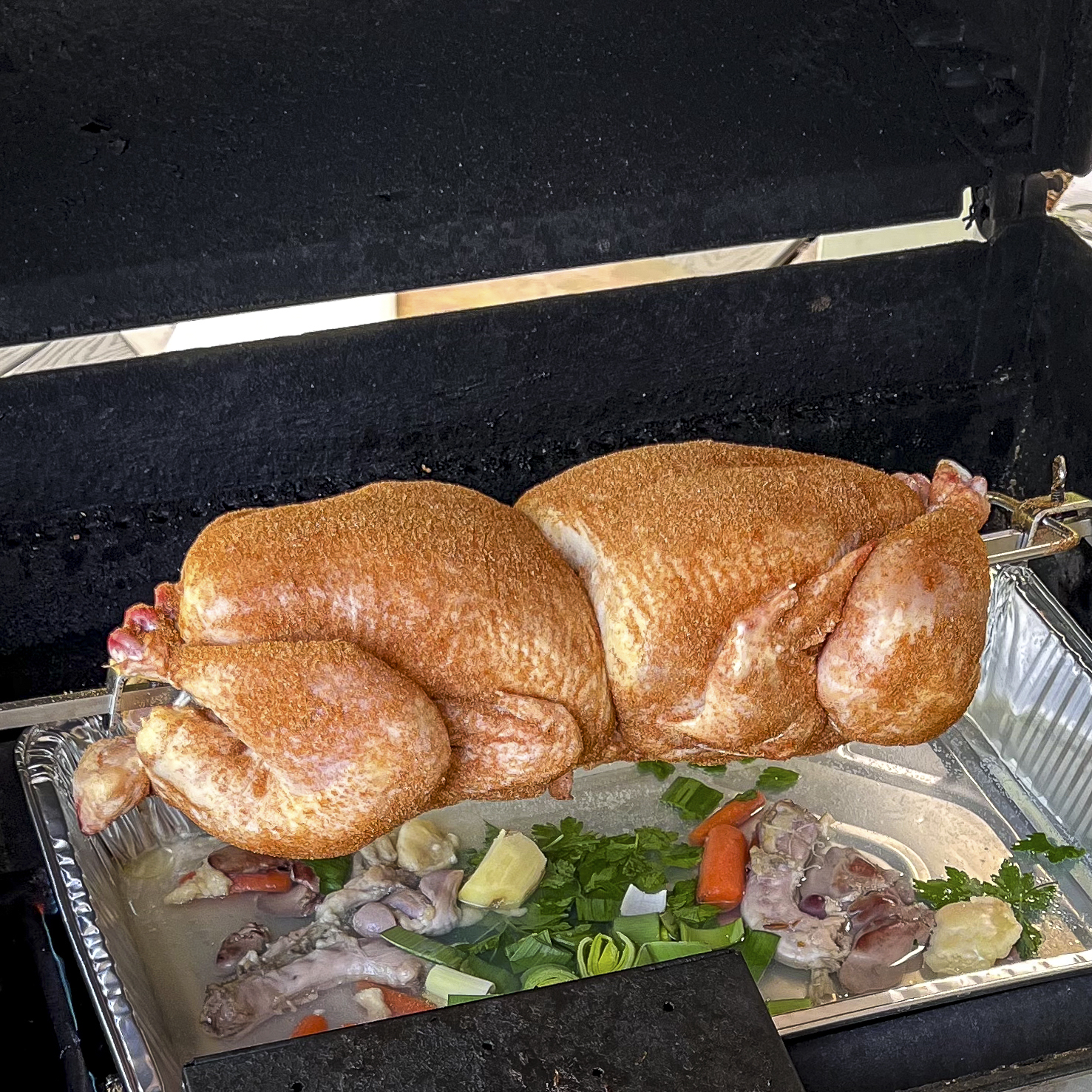 Two seasoned chickens are in the grill on a rotisserie spit rod.