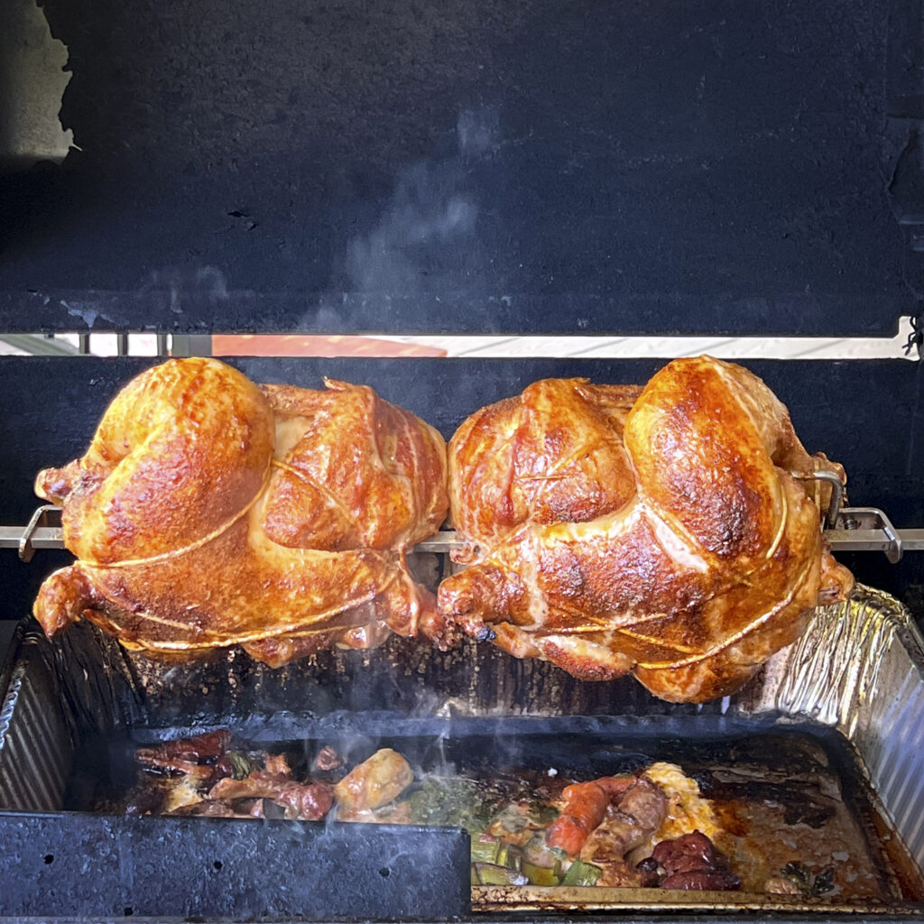 Two chickens are almost done cooking on a rotisserie in a gas grill. 