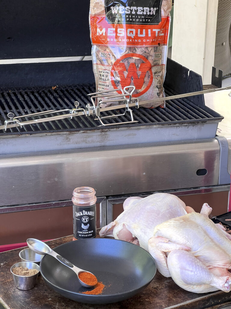 A bag of Mesquite Smoking Chips is on a grill with a spit rod to show what happens next. A chicken and seasoning is in front of the photo. 
