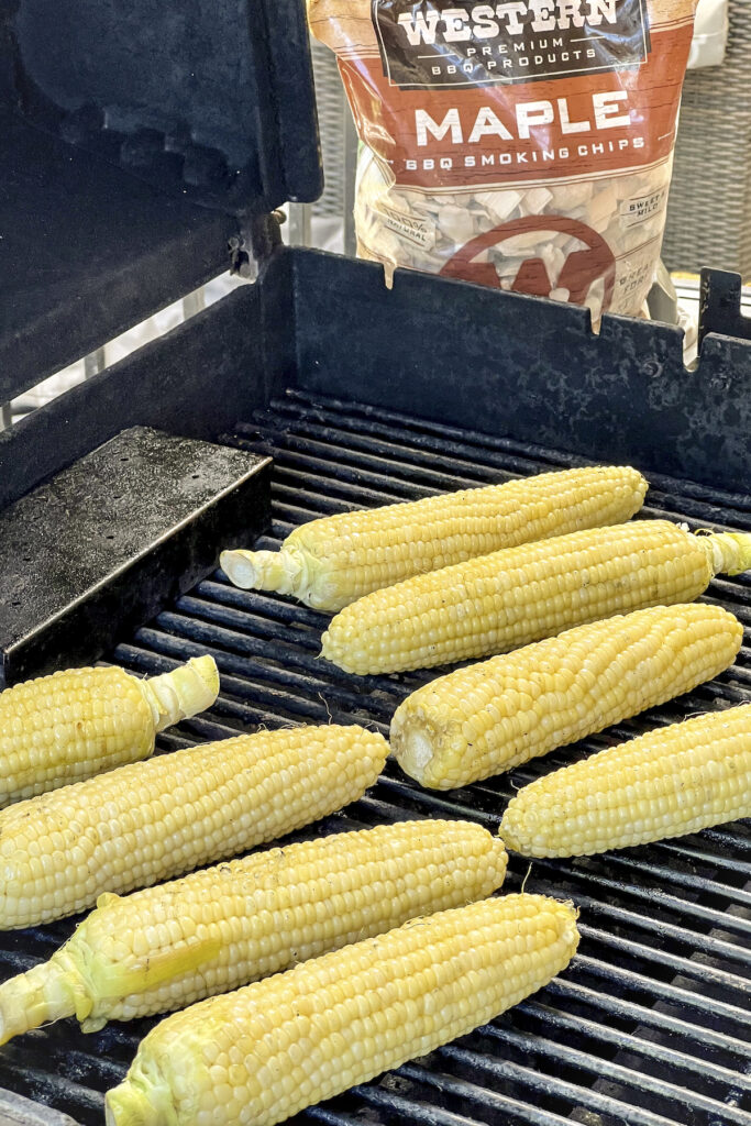A smoker box is on the grill. Eight blanched corn on the grill with Maple Smoking Chips near by.
