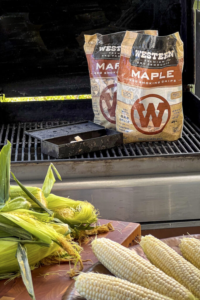 Western BBQ Maple Smoking Chips on a grill next to a smoker box. Up front there are ears of corn that have not been shucked. 