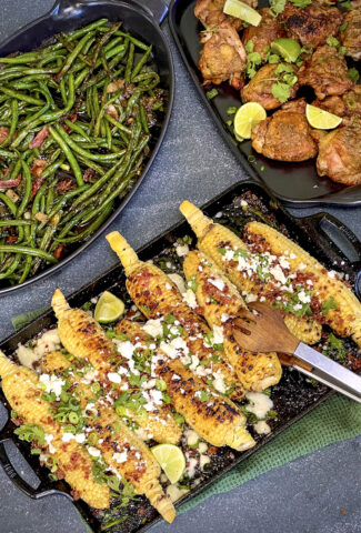 Smoked, maple glazed, and grilled corn is topped with cheese, scallions, and cilantro and is ready to be served. Green beans and chicken are in the photo to show what to serve with the corn.