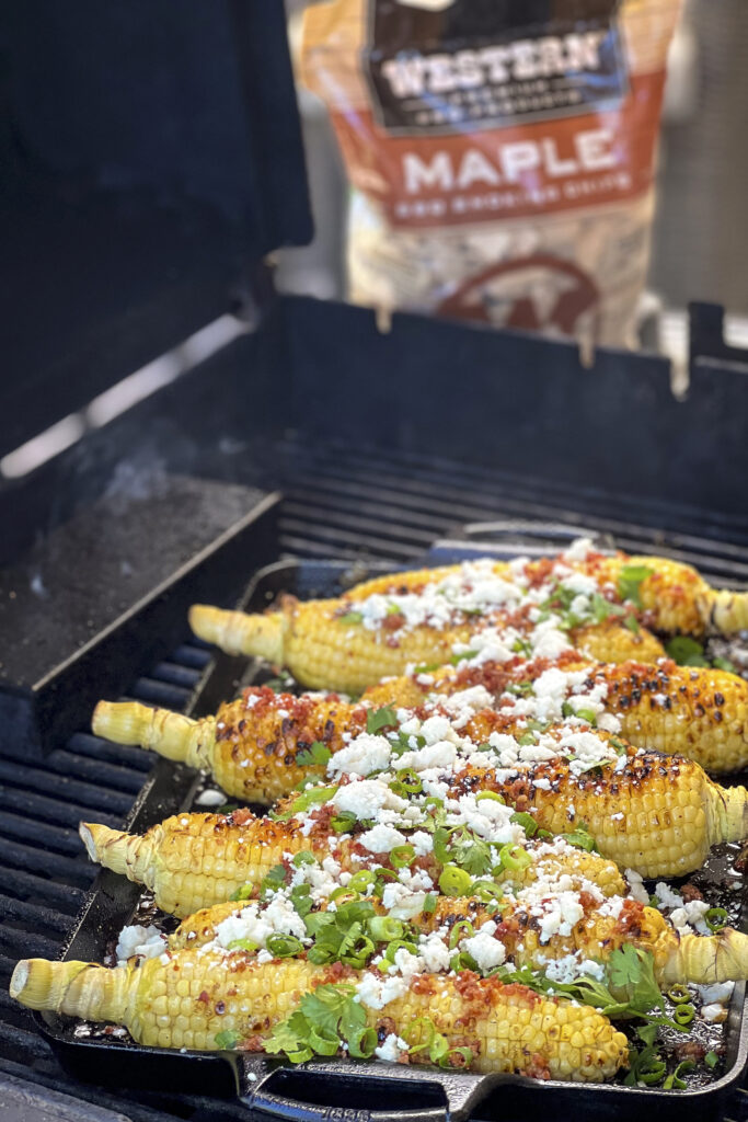 Smoky corn is on the grill topped with queso fresco, bacon bits and cilantro.  