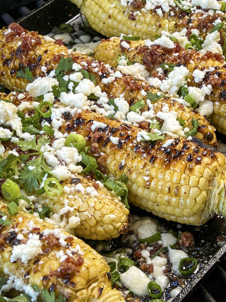 A close up photo of maple smoked corn on the cob with queso fresco, scallions, cilantro, and bacon bits. 