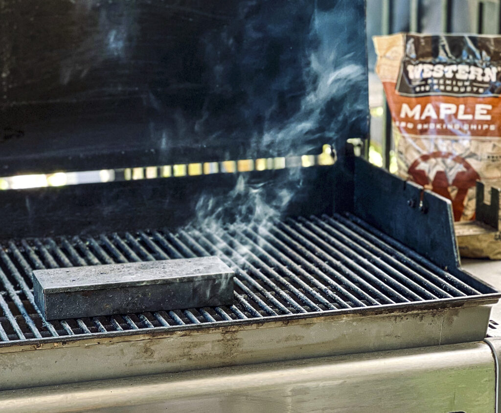 A grill is open with a smoker box that has smoke drifting from it. A bag of Western BBQ Smoking Chips is in the background.