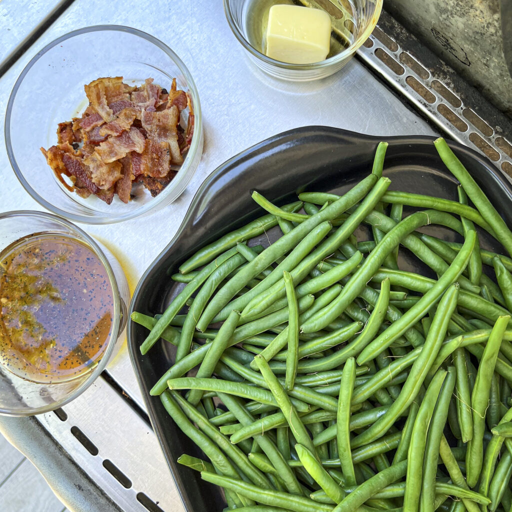 A black serving bowl of blanched green beans waiting to be taken to grill. Bacon, butter, and a maple glaze are on counter too.