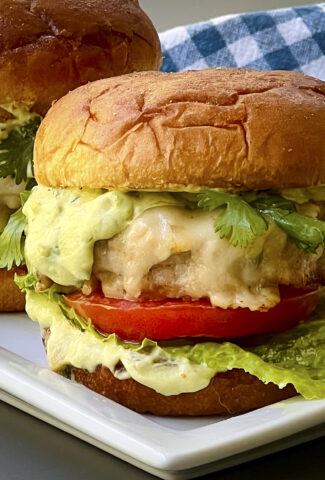 A chicken burger smoothered in melted cheese tucked in a golden bun. A slice of tomato and a leaf of lettuce on on the bottom bun. Avocado cream is dripping over the ground chicken burger.