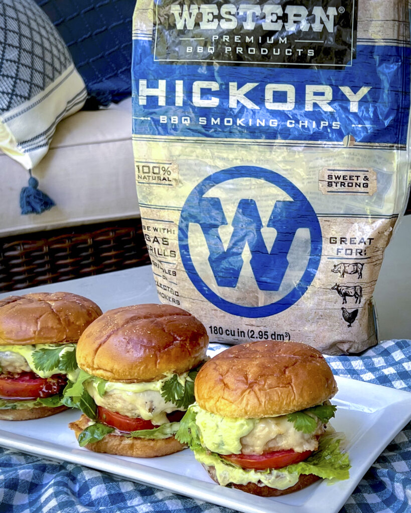 A bag of Western Hickory Chips is in the backdrop with 3 chicken burger in buns with garnishes. 