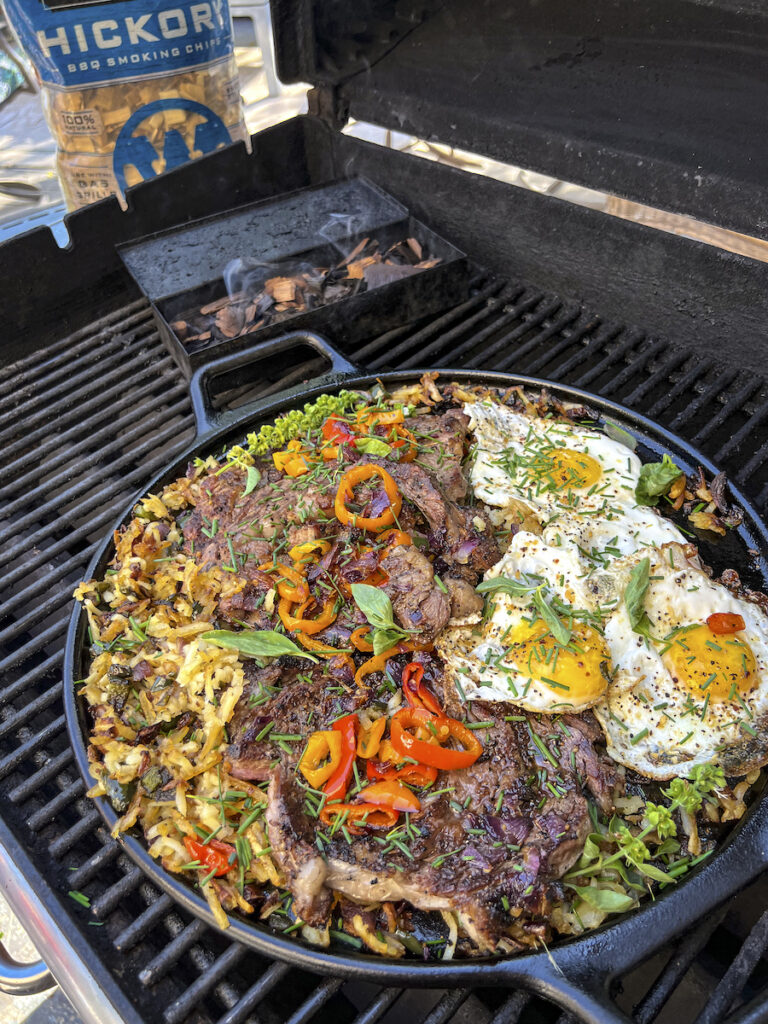 A cast iron skillet has smoked and grilled hash browns, rib eye steaks and eggs.