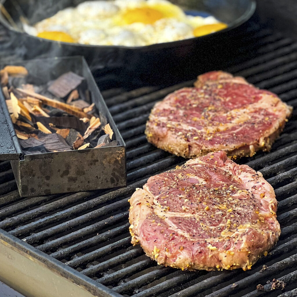 2 ribeye steaks with smoker box and eggs in the back drop. 