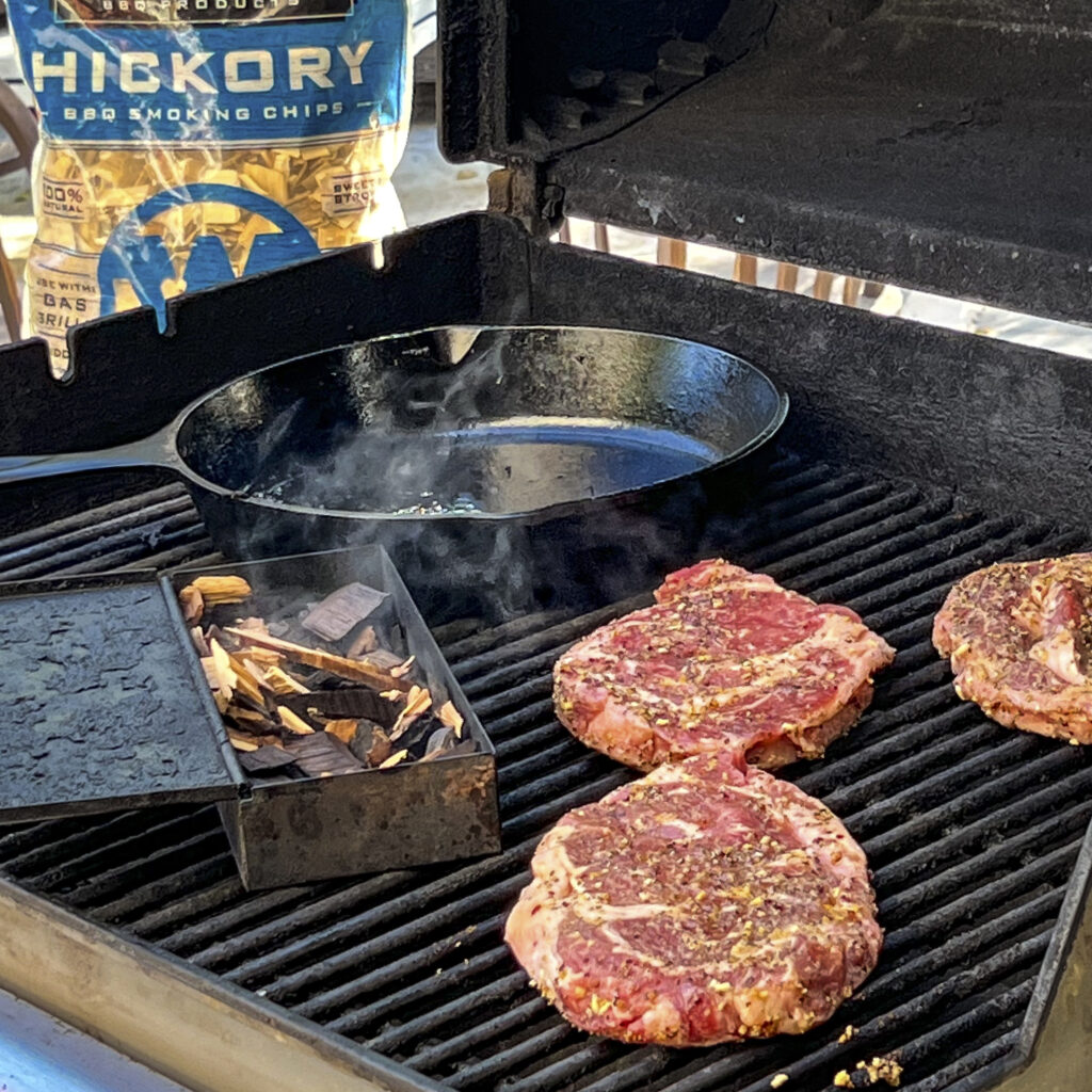 A grill shot with ribeye steaks on it, a smoking box, and a cast iron skillet. A bag of Western BBQ Smoking Chips is in the backdrop. 