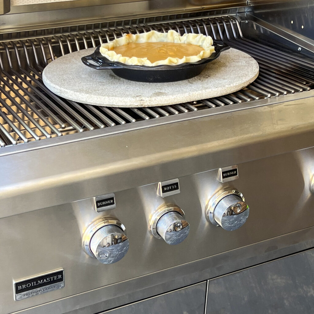 A pumpkin pie sits on a baking stone in a grill.