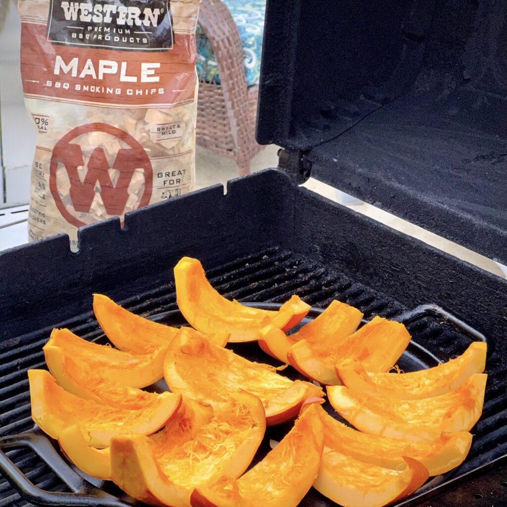 Wedges of pumpkin are on a grill smoking. A bag of Western BBQ Smokin Chips is in the background.