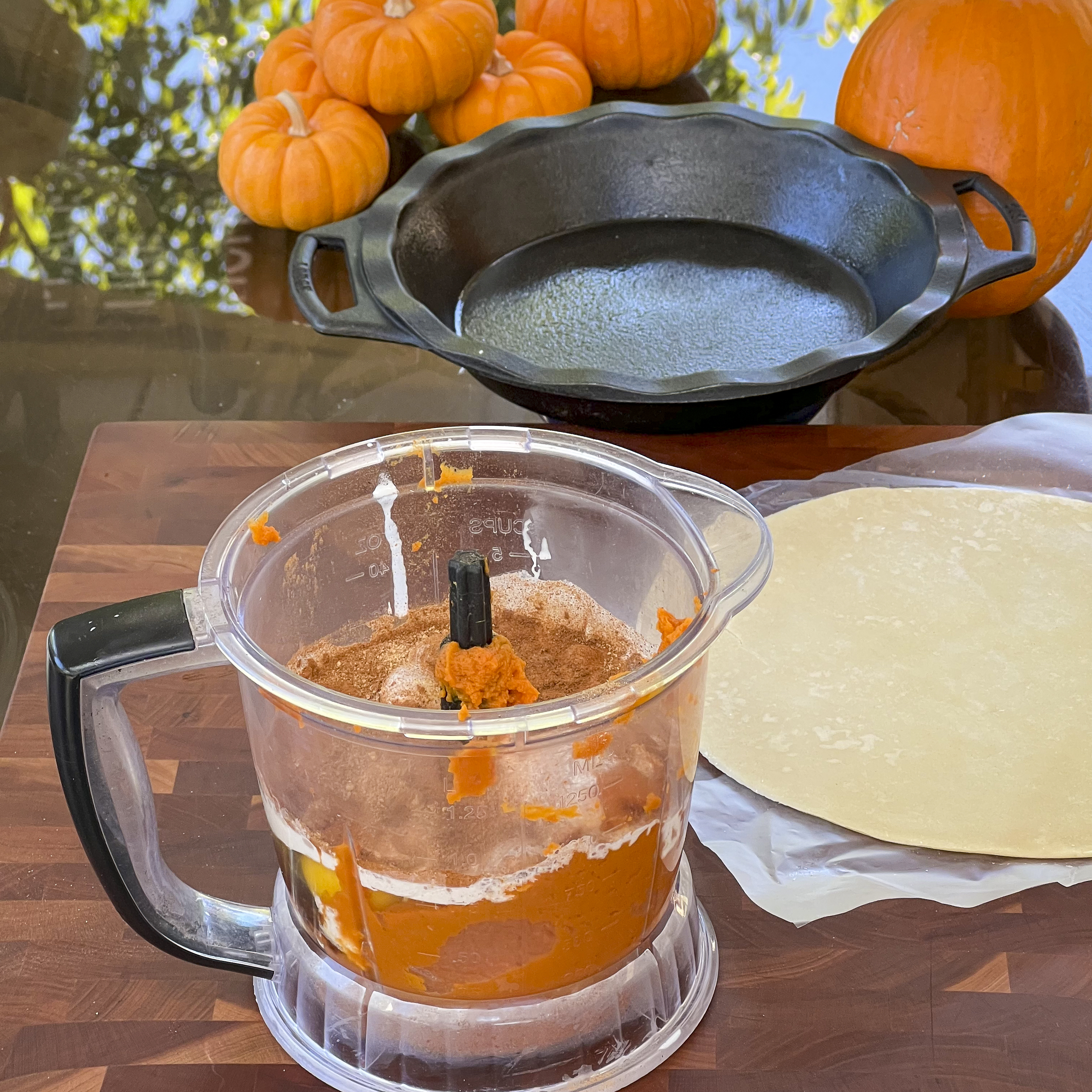 A food processor holds pumpkin puree and other ingredients ready to blend. Near by is a pie dough round and a cast iron pie pan. 