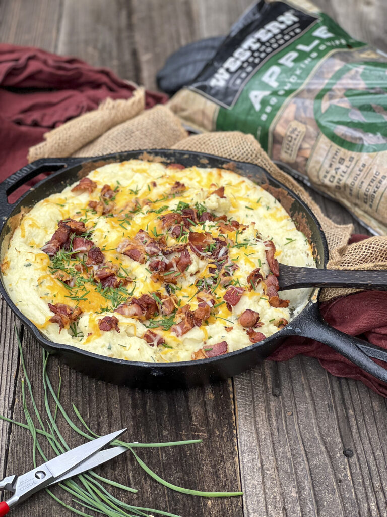 A cast iron skillet is in focus with chives and a pair of scissors. In the backdrop an out of focus bag of Western BBQ Apple Smoking Chips.