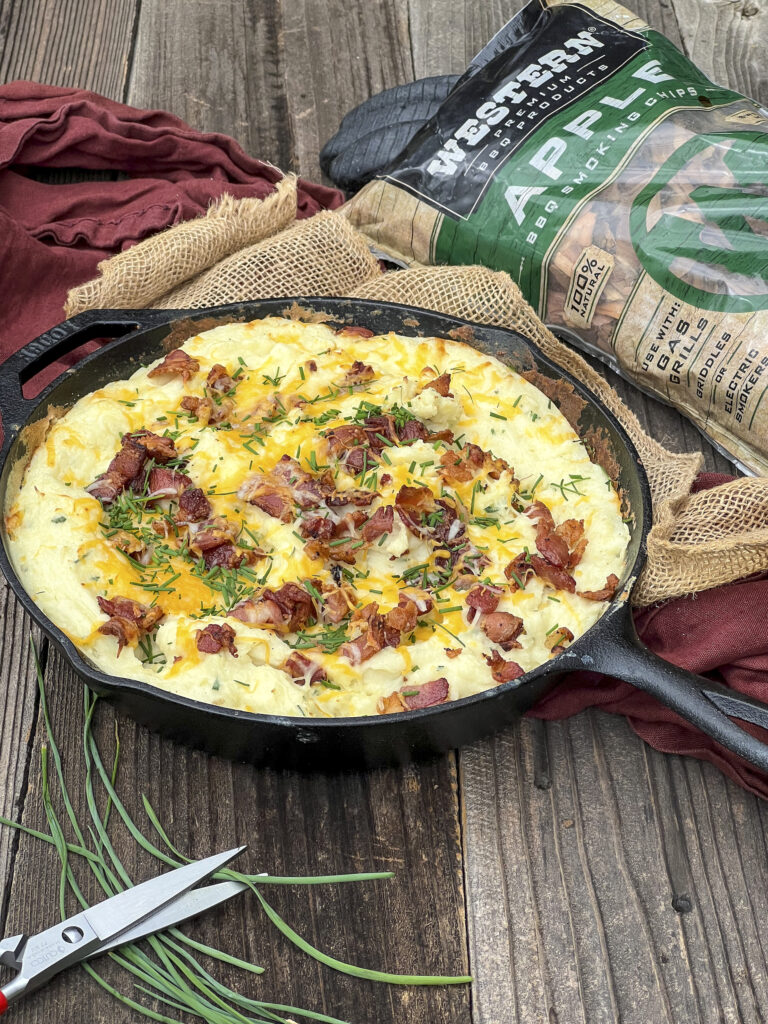 Cast iron skillet has smoked mashed potatoes with bacon, chives, and cheese. A bag of Western BBQ apple chips is in focus too.