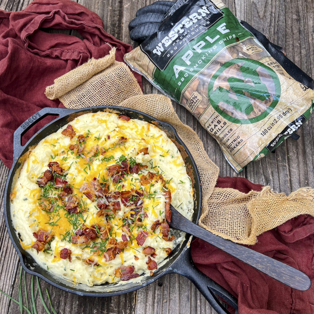 An overhead of a cast iron skillet that a holds mashed potatoes with cheese, chives, and bacon on top. There is a bag of Western BBQ Apple Smoking Chips near by to show what smoke was used.