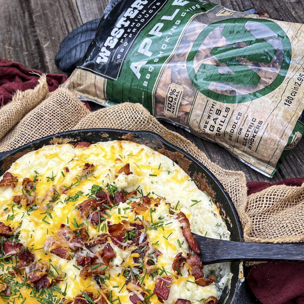 A bag of Western Apple Chips is near by a cast iron skillet of smoked mash potatoes with bacon crumbles, chive and cheese.