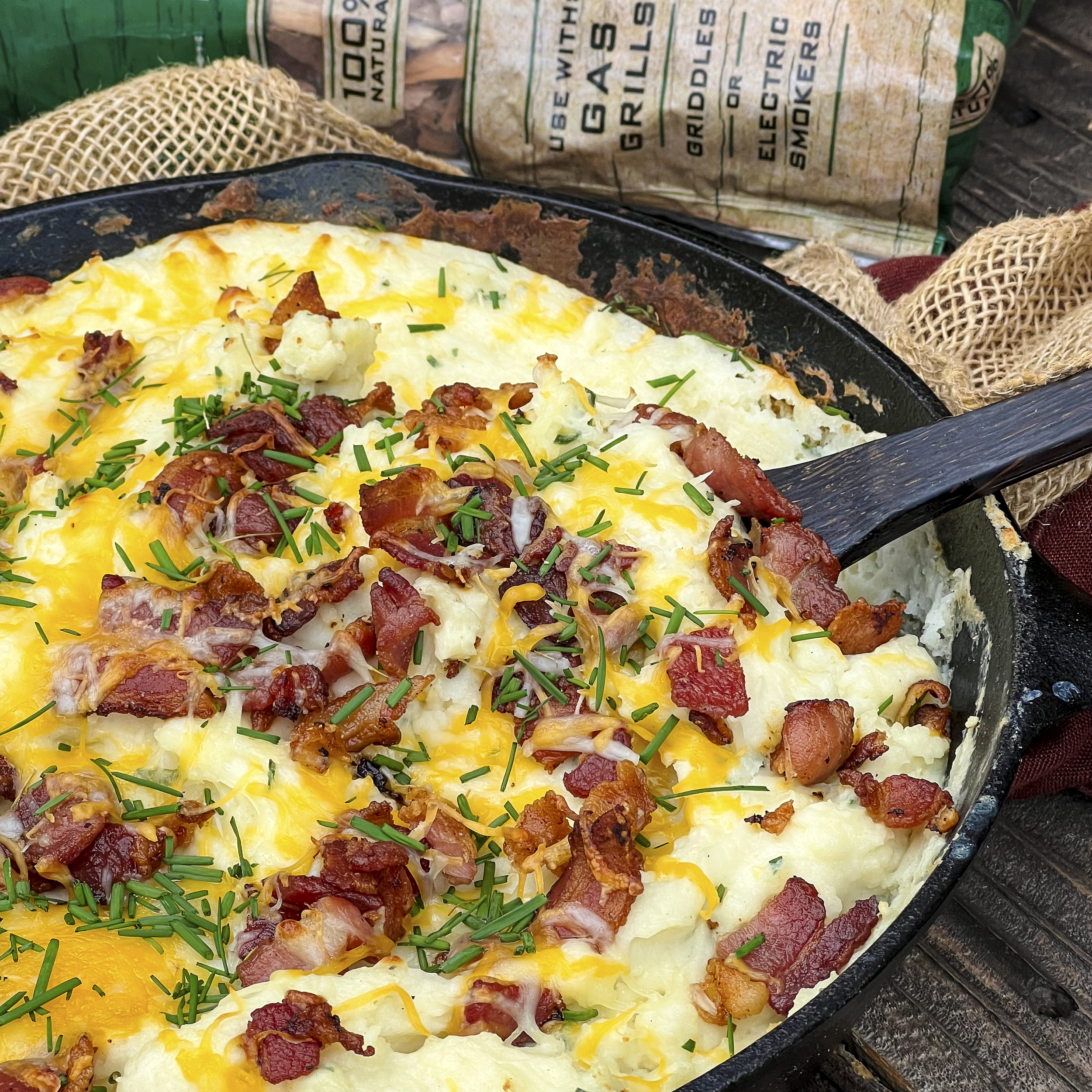 A cast iron skillet with mashed potatoes and big bacon pieces, chives, and shredded cheddar cheese.