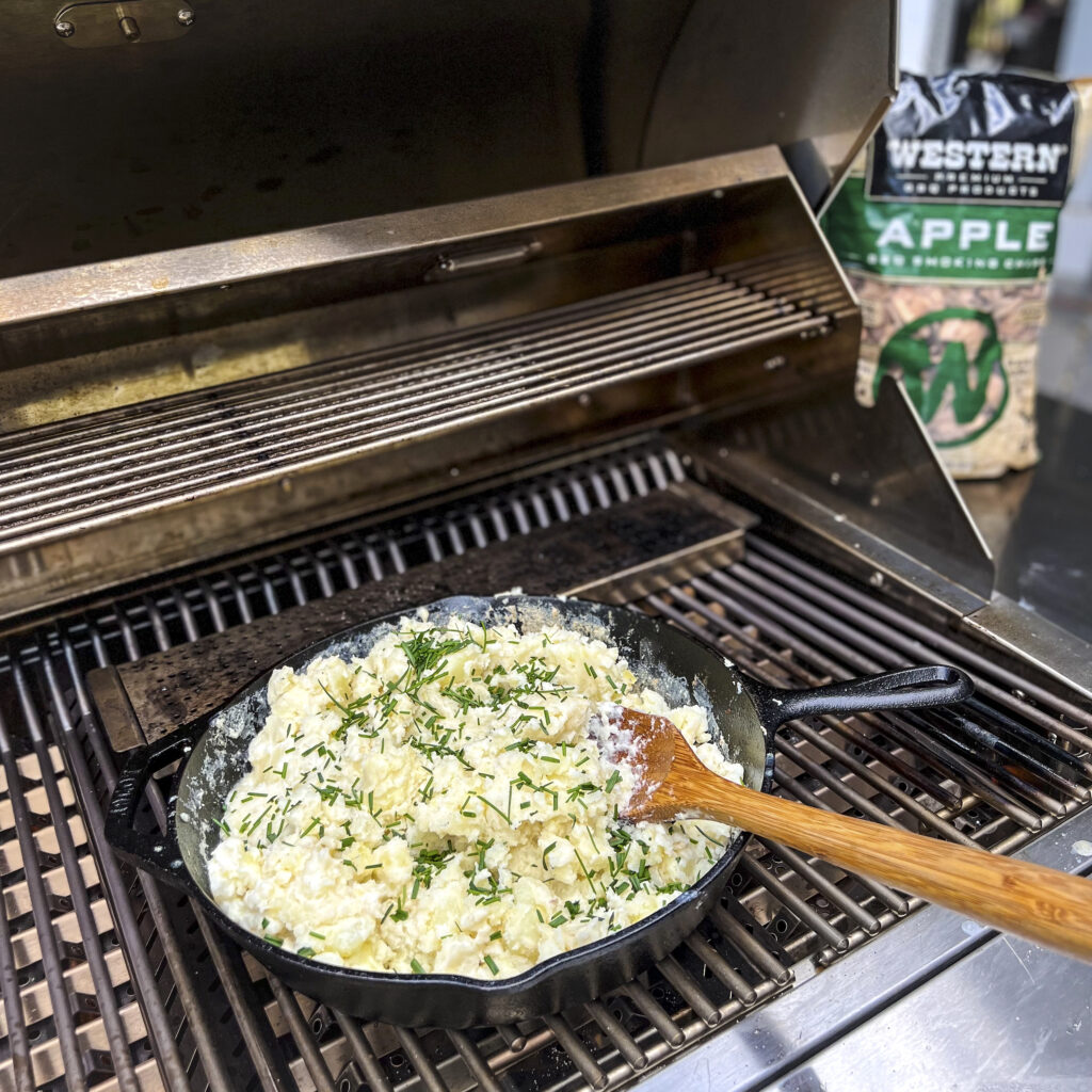 A cast iron skillet is on the grill with a mound of mashed potatoes that have chives sprinkled on top.