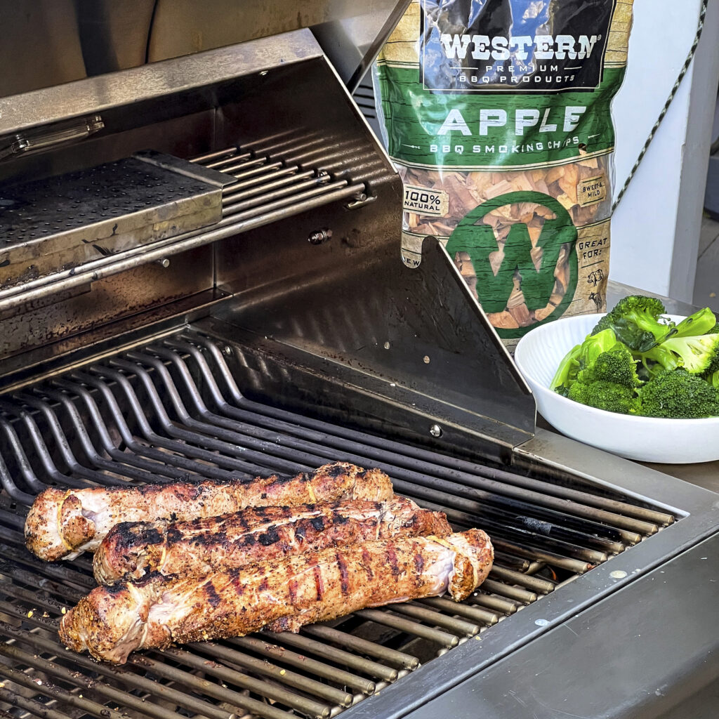 Pork Tenderloin is on the grill with a bag of Apple Smoking Chips and Broccoli in the backdrop. 