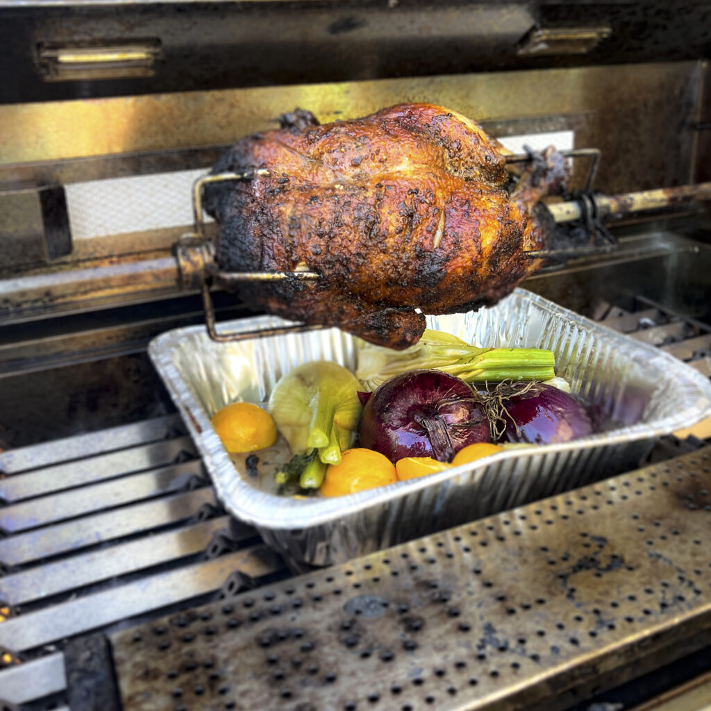 Duck is on a rotisserie with a drip pan that is filled with aromatics.