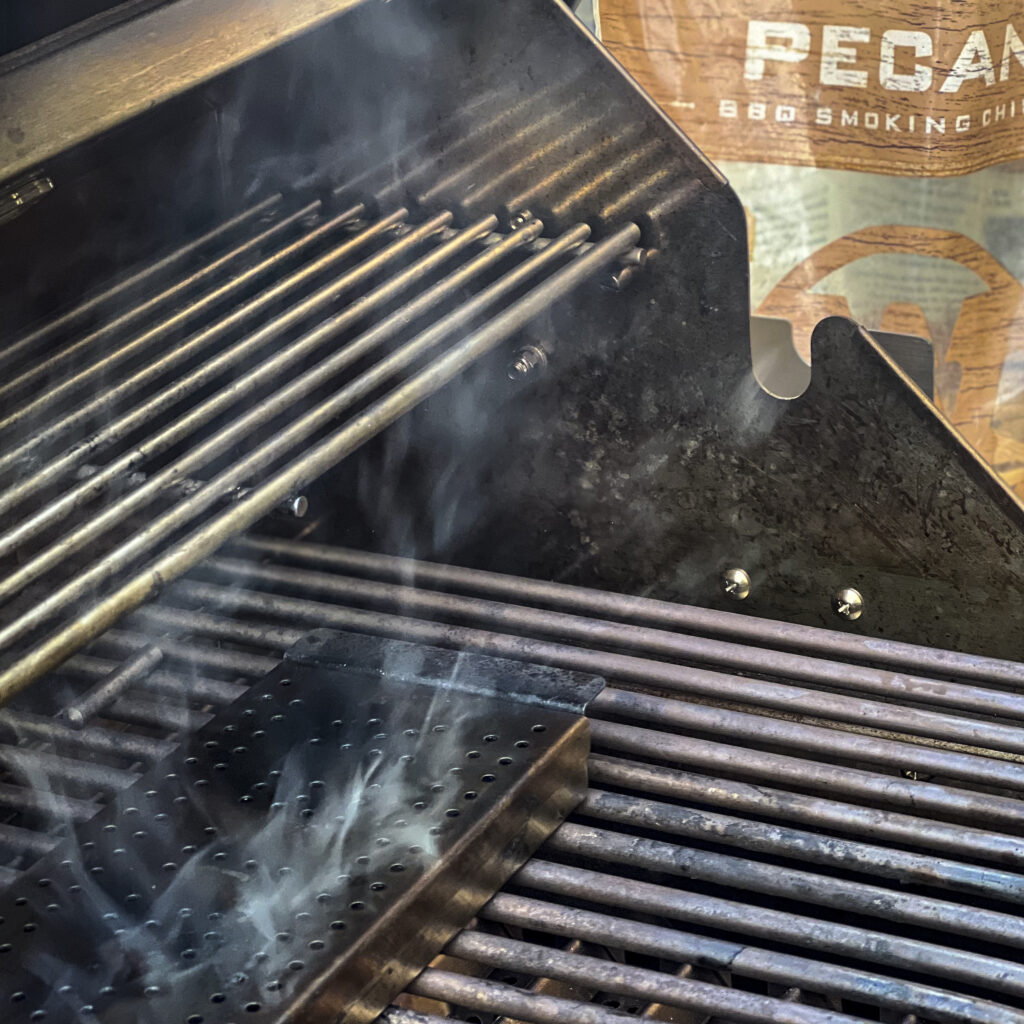 A smoker box with smoke drifting from it on a gas grill. In the backdrop there is a bag of Western BBQ Pecan Smoking Chips. 
