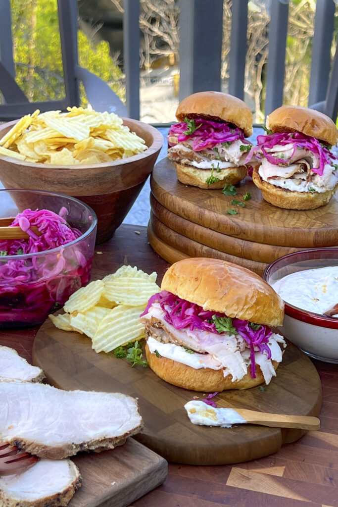 Hickory Smoked Pork Loin Sandwiches on wood plates with potato chips. The pork slices, on a brioche bun, and the pickled cabbage, parsley, and horseradish spread make a bright scene.