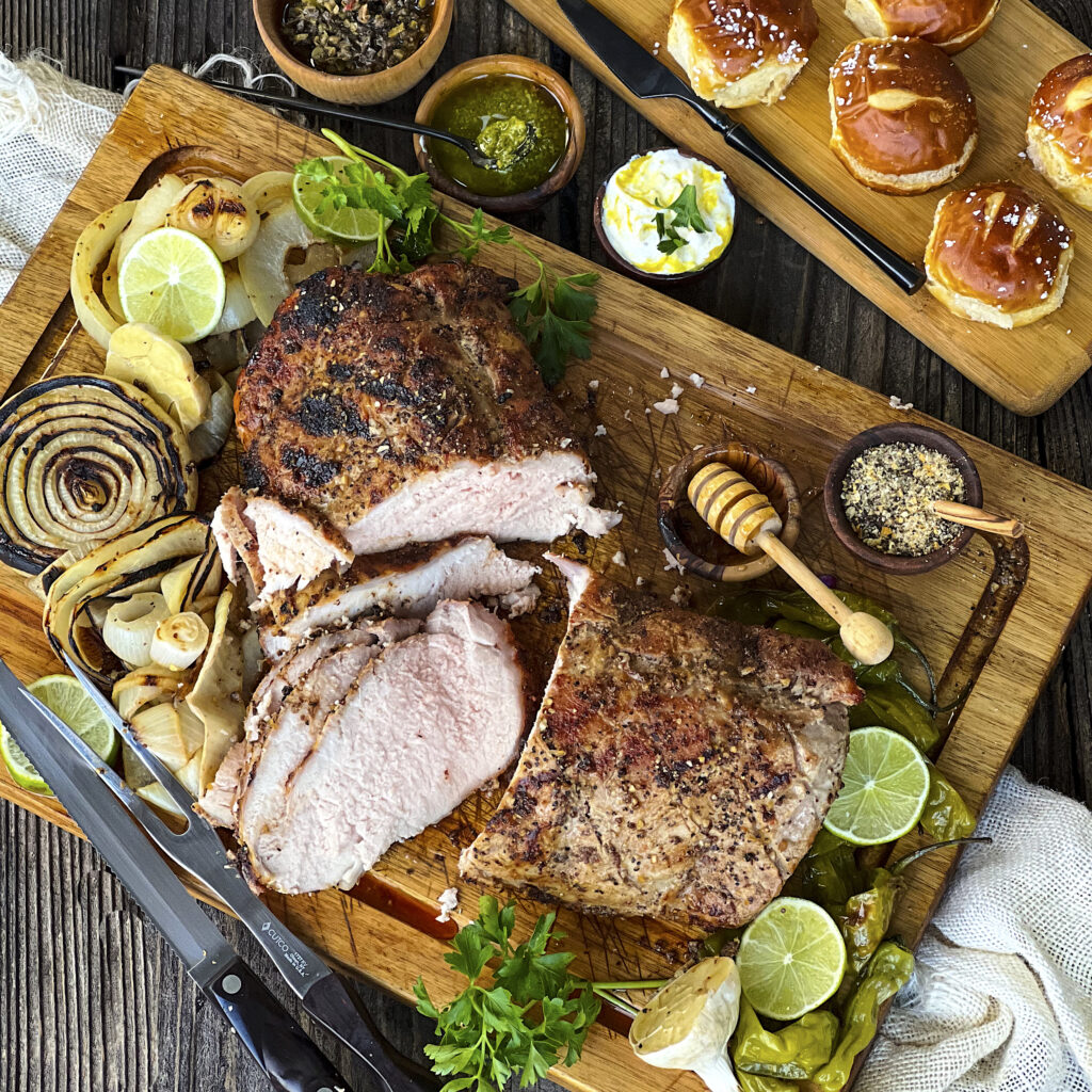 Hickory Smoked Pork Loin is sliced on a cutting board with pretzel buns in the corner. 