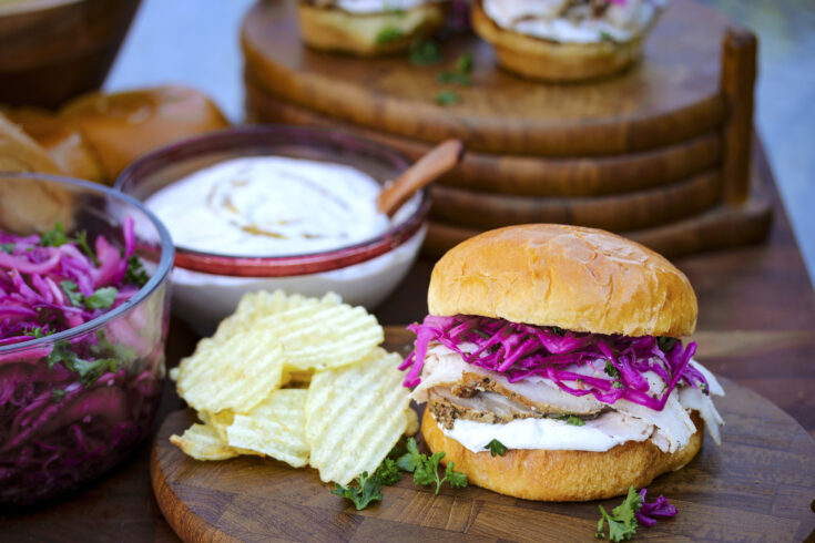 A close up of a pork loin sandwich with pickled red cabbage and onions, plated with ruffled potato chips.