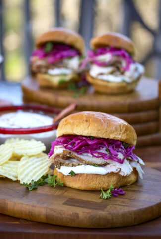 3 hickory smoked pork loin sandwiches with pickled red cabbage and a creamy horseradish sandwich spread. Potato chips are being served on the wood plate with the brioche sandwich.