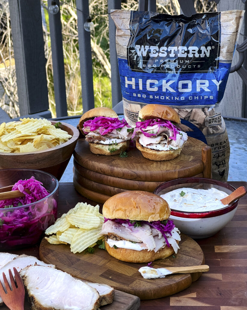 A bag of Western Hickory BBQ Smoking Chips are in the background of a smoked pork loin sandwich scene. Potato chips are being served. The brioche buns are filled with smoked pork loin, pickled red cabbage, and horseradish spread.