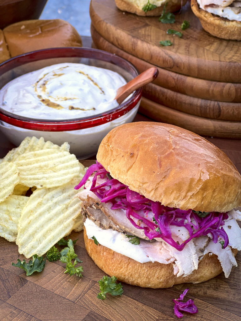 A close up of one pork loin sandwich with pickled red cabbage and onions. Potato chips are being served along with a homemade horseradish sauce.