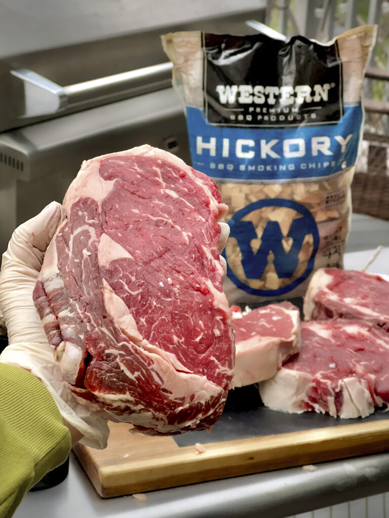 A ribeye steak is in the front with a Western BBQ Hickory Smoking Chips in the back. 