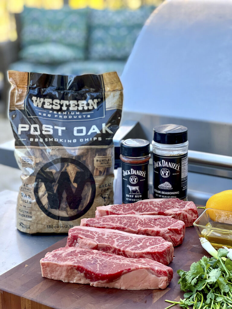 A cutting board with 4 strip steaks and Jack Daniel's Seasoning are in the scene. A bag of Western Post Oak Smoking Chips has been included.