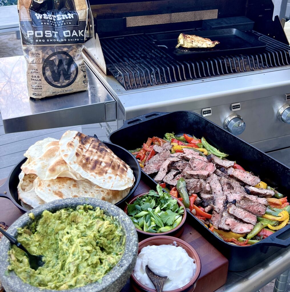 A grill scene with sliced smoked steaks, guacamole, tortillas, sour cream and cilantro.