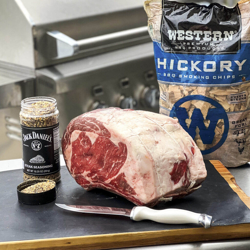 A whole bone-in ribeye roast is on a cutting board with Jack Daniel's Seasoning and Hickory Smoking Chips. 