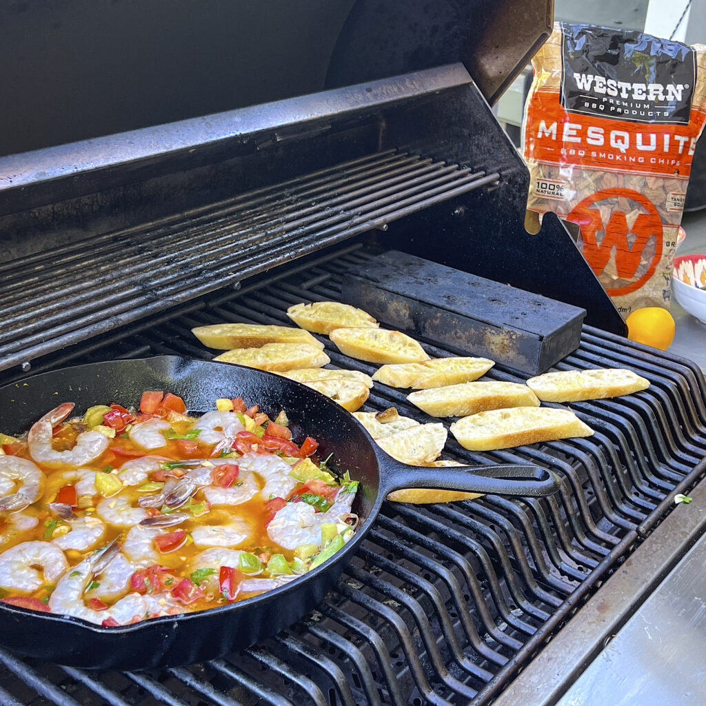 Slices of a baguette are on the grill to toast. A skillet of tomato butter sauce is in the skillet. A bag of Mesquite Smoking Chips is near by. 