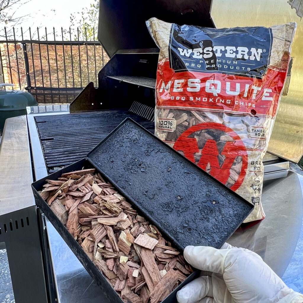 A smoker box with Western BBQ Mesquite Smoking Chips is near the grill with a box full of the smoking chips. 