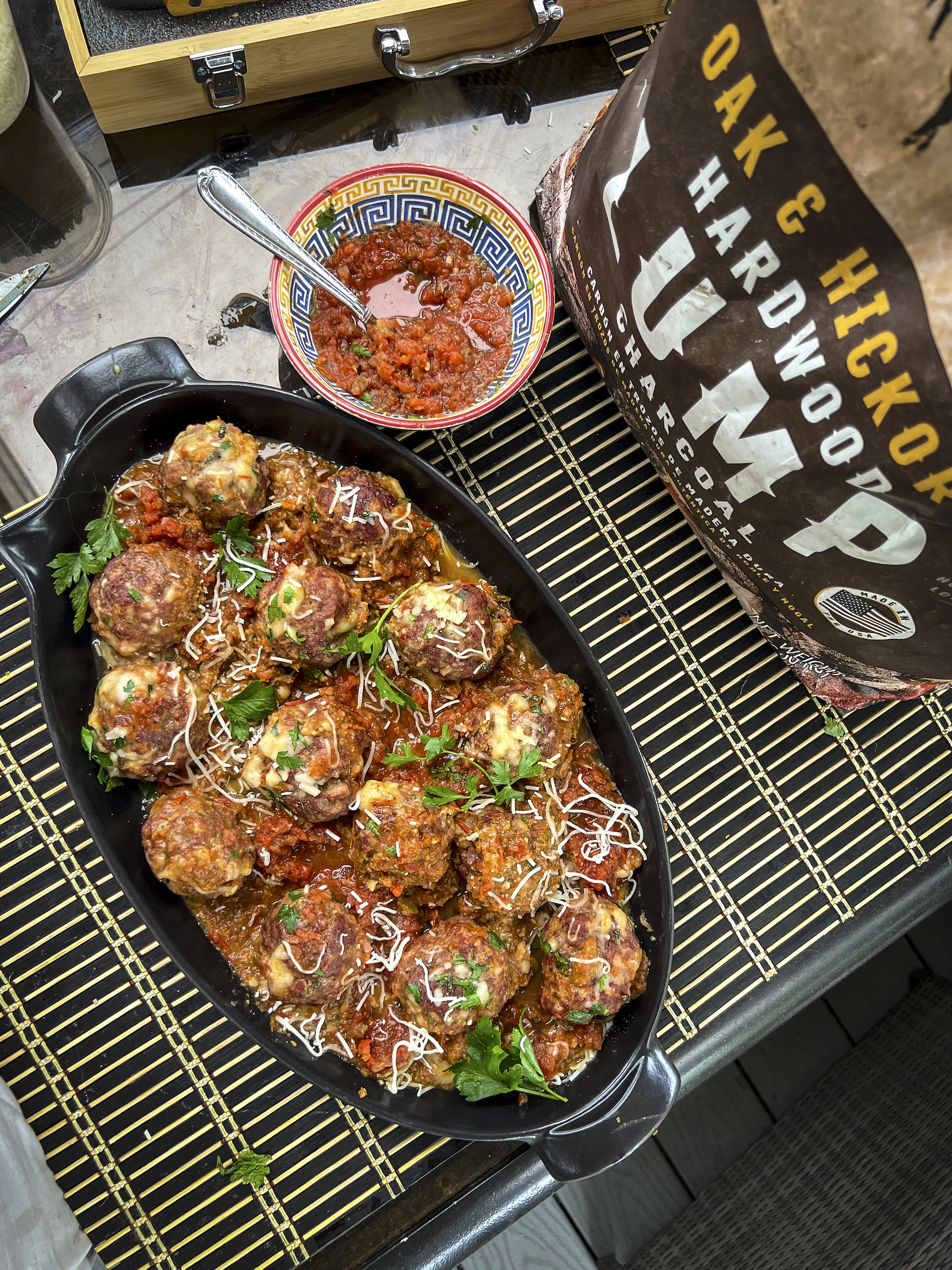 Smoked Meatballs are in a black serving dish. There is a partial bag of Lump Charcoal and a bowl of pasta sauce.
