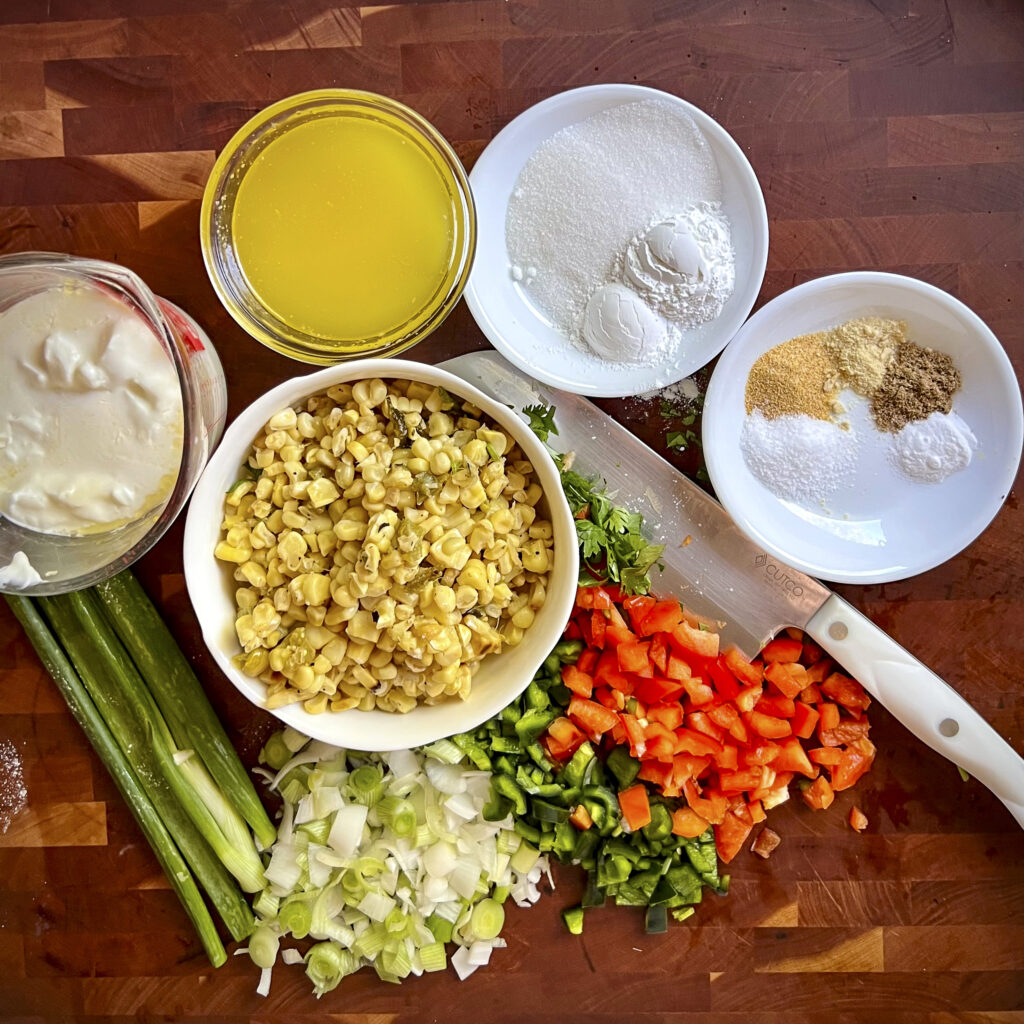 A photo with grilled corn kernels in a bowl, sugar, baking soda and powder on a white plate. There is a small white plate with salt, coriander, garlic, and onion powder. In the photo there is chopped onion, poblano, and red bell pepper.
