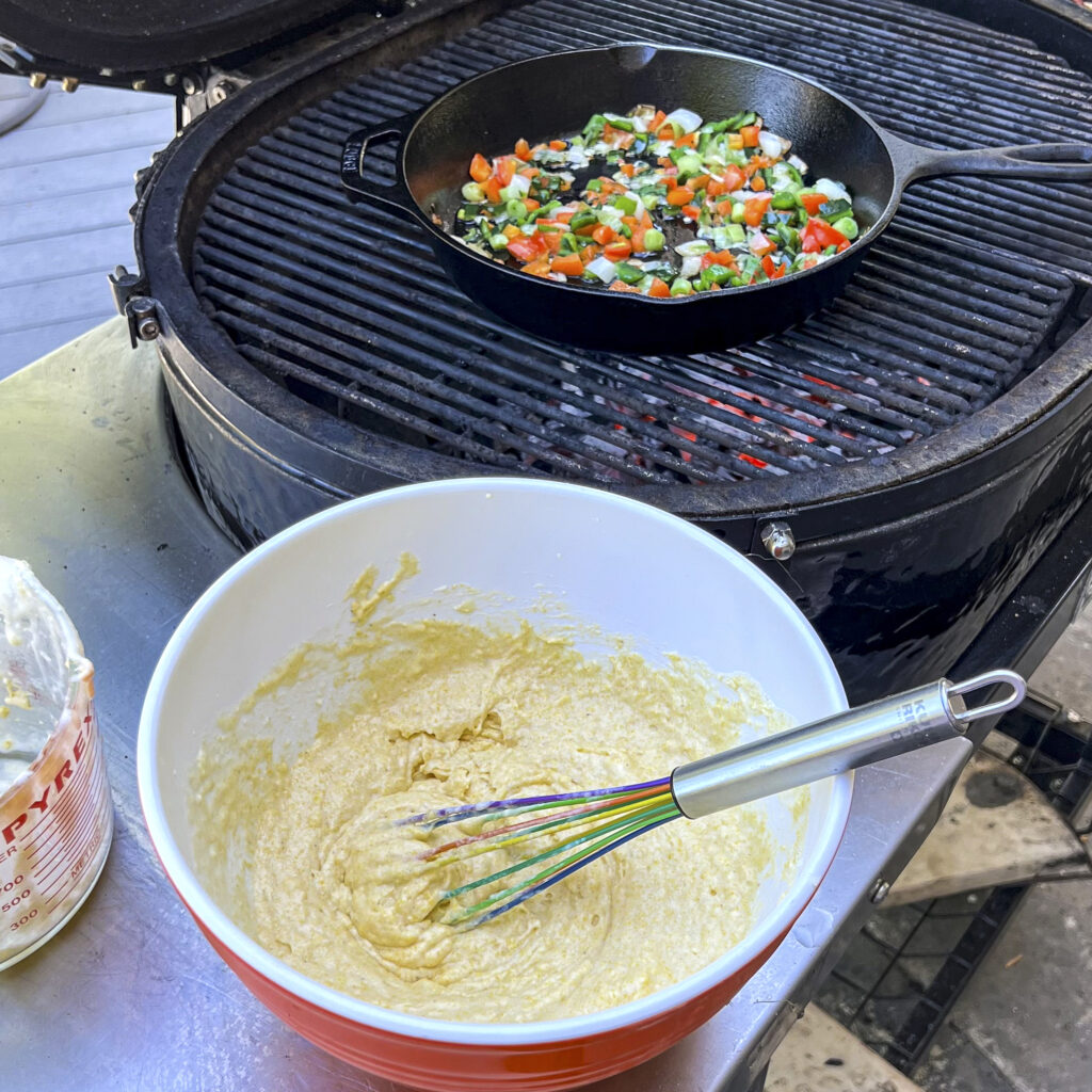 A bowl of cornbread batter is in the front. There is a skillet with peppers and onions, on the grill, in the background.